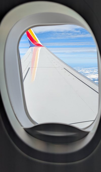 A view through an airplane window shows the wing with Southwest Airlines' red, yellow, and blue colors against a partly cloudy sky.