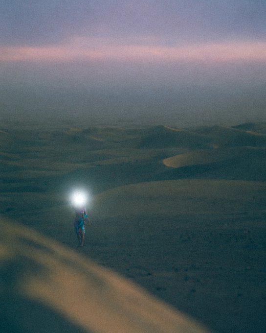 A solitary figure wearing a headlamp walks through vast sand dunes at dusk, creating a glowing orb of light against the muted, atmospheric desert landscape.