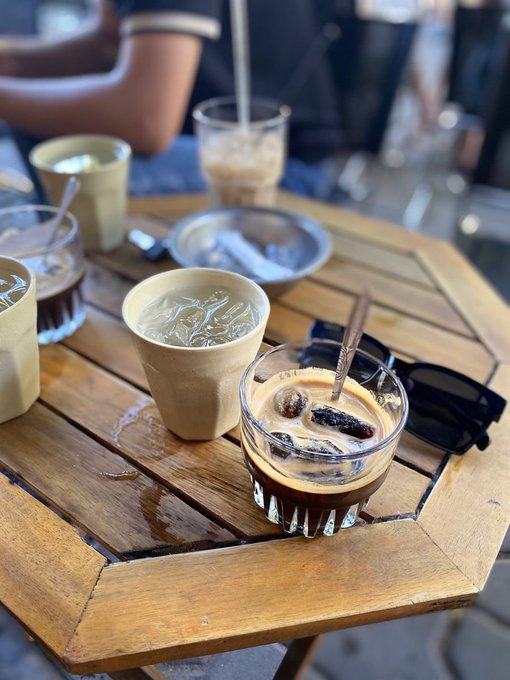 A wooden table at an outdoor cafe holding several glasses and cups of iced coffee drinks, with a pair of black sunglasses resting on the table.