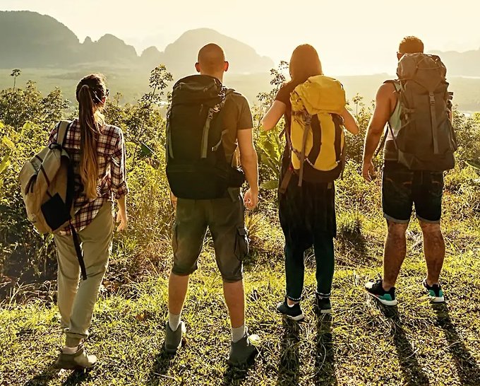 Four hikers with backpacks stand on a grassy hillside at sunset, looking out toward distant mountains and a valley below.