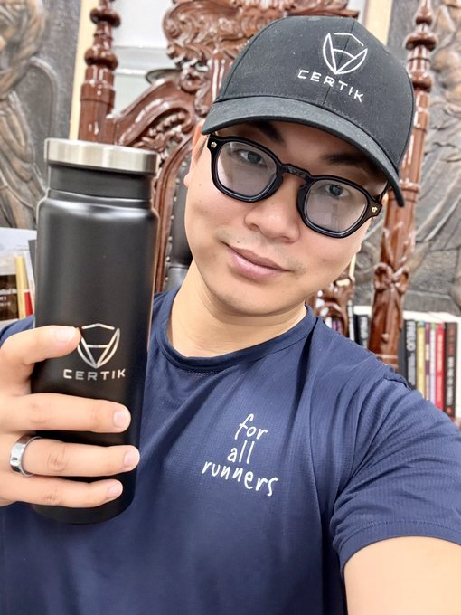 A selfie of a person wearing a navy 'for all runners' t-shirt, black glasses, and a black 'CERTIK' branded cap, holding a black 'CERTIK' branded water bottle.