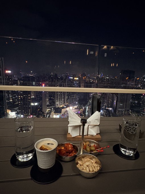 A rooftop dining setup at night featuring two glasses of water, a cup of coffee, small bowls of snacks, and a napkin holder, overlooking a sprawling, illuminated city skyline.