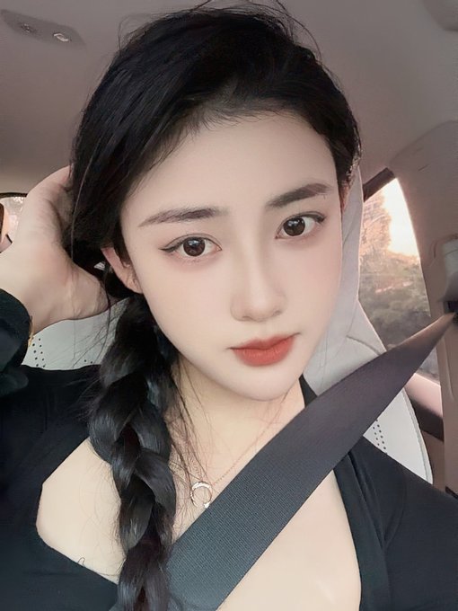 A selfie taken inside a car shows a woman with a long dark braid wearing a black top and a red lip, secured by a seatbelt.