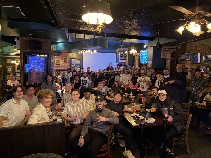 A large group of people posing for a photo inside a traditional-style pub, with warm lighting from chandeliers and a TV screen in the background.