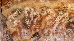 An ancient cave painting featuring numerous red handprints arranged on a textured rock surface.