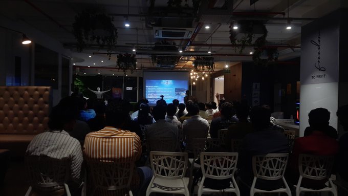 A dimly lit presentation or meetup event where attendees sit in chairs facing a projection screen, with a speaker gesturing on stage and a wall sign reading 'TO WHITE'.
