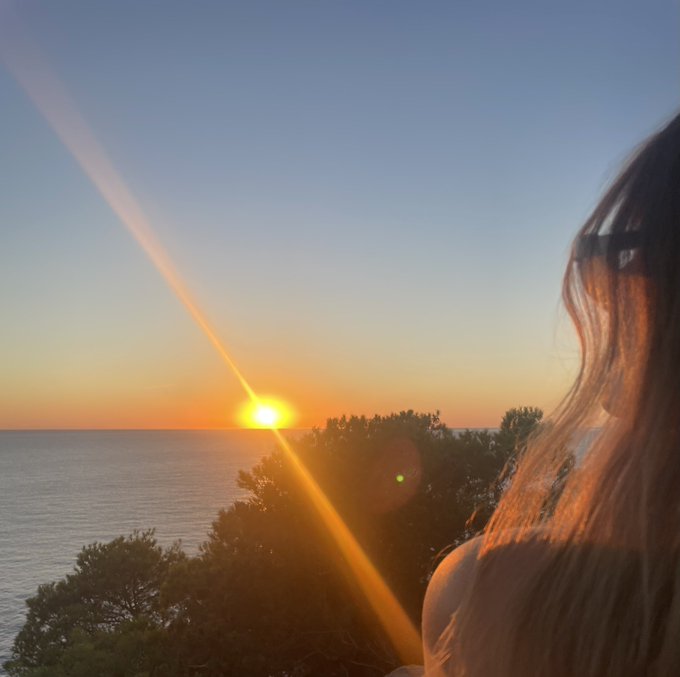 A scenic photo captures a bright sunset over the ocean with trees in the foreground, while a woman with long dark hair and sunglasses is partially visible on the right side of the frame.