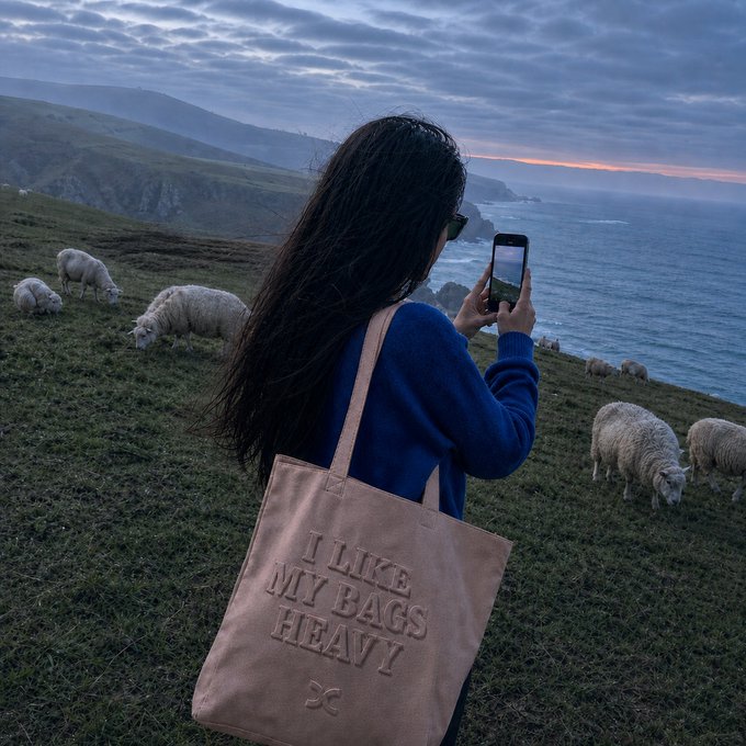 A woman takes a photo of sheep grazing on a coastal cliff at dusk, carrying a tote bag that reads "I LIKE MY BAGS HEAVY," humorously echoing the "touch grass before touching leverage" message by suggesting she prefers tangible, heavy belongings over risky financial bets.