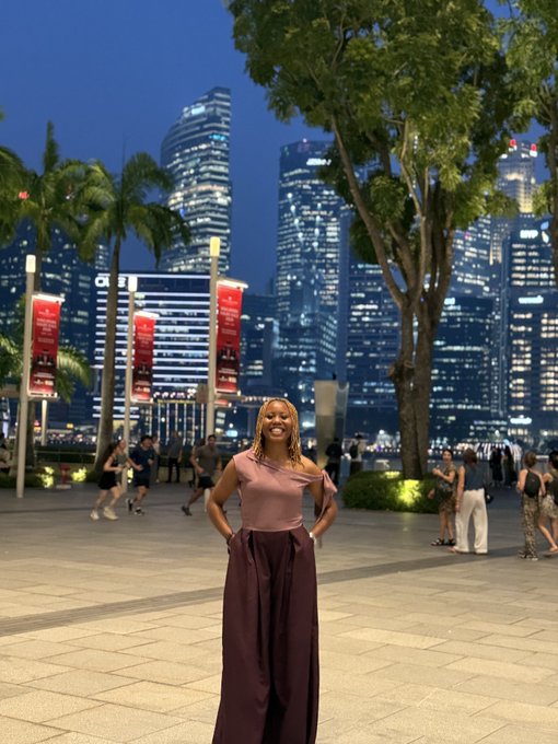 A woman poses confidently with her hands on her hips in front of the illuminated Singapore skyline at night, with palm trees and banners lining the waterfront promenade behind her.