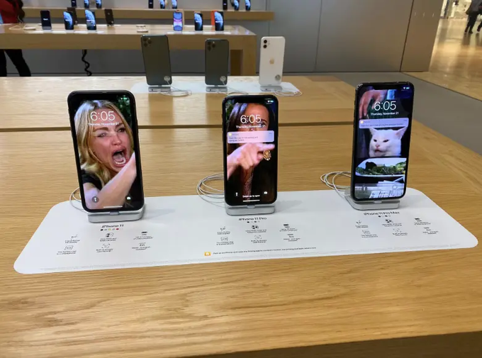 Three iPhones are displayed on wooden stands inside an Apple Store, showing lock screens with the time 6:05. The labels below identify them as 'iPhone 11', 'iPhone 11 Pro', and 'iPhone 11 Pro Max'.