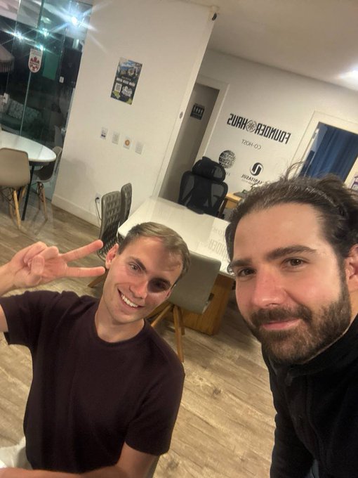 Two men taking a selfie in what appears to be an office space, with one making a peace sign and the other with a beard; the word 'LONGERIZE' is visible on the wall behind them.