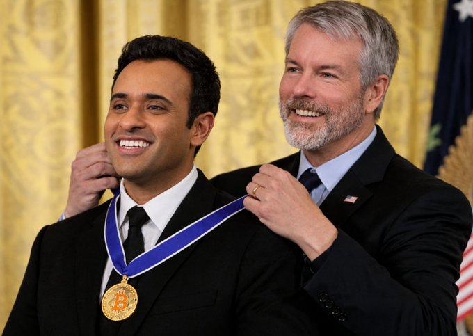 Two men in suits in a formal setting with gold curtains and an American flag, as one man places a blue-ribboned medal around the other's neck.