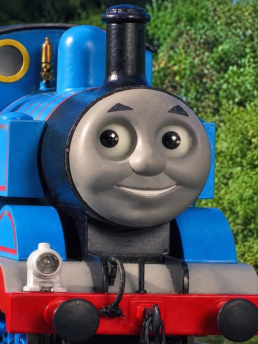 A close-up of Thomas the Tank Engine, the blue steam locomotive character with a smiling face, set against a background of green foliage.