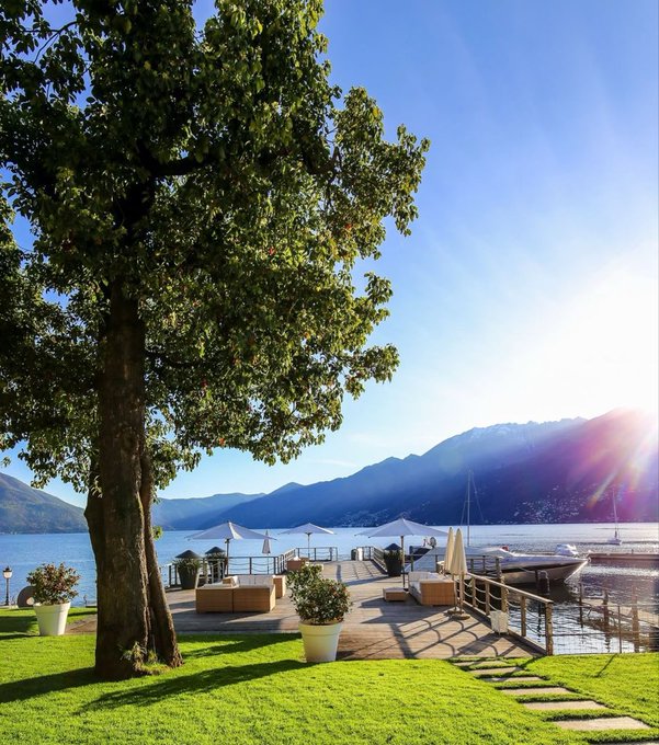 A scenic lakeside view featuring a large tree in the foreground, a wooden deck with white umbrellas, potted plants, and boats docked at a pier, with mountains and a bright sun in the background.