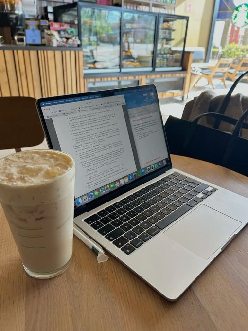 A Starbucks iced coffee next to an open MacBook showing a website, with the Starbucks storefront visible in the background.