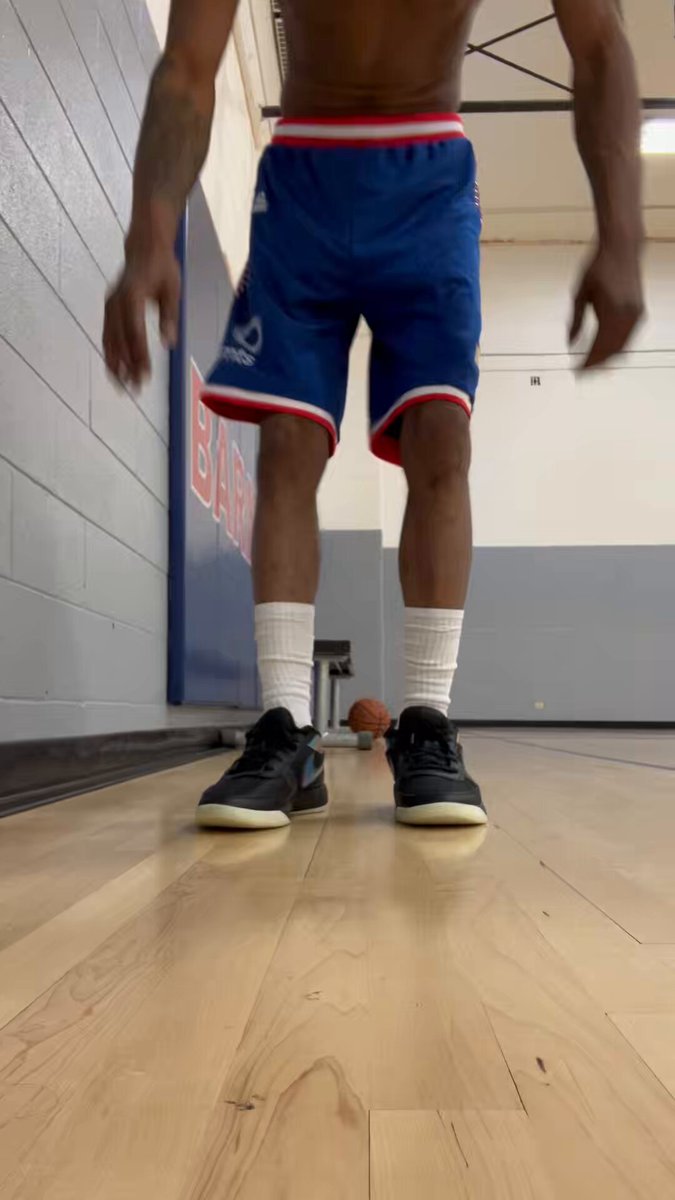 The image shows a man wearing blue basketball shorts and black shoes standing on an indoor basketball court, with his arms outstretched as if he's about to jump or perform a trick. The floor of the gym has a wooden surface that matches the color of the man's shorts. There is also a basketball visible in the background, suggesting that this scene takes place at a basketball facility.