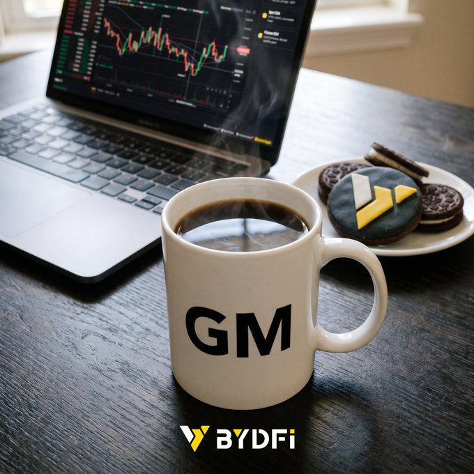 A steaming mug labeled "GM" sits beside a laptop displaying trading charts and a plate of cookies, embodying the "gm" greeting with a cozy twist on the classic crypto ritual of starting the day with coffee and chart-watching.