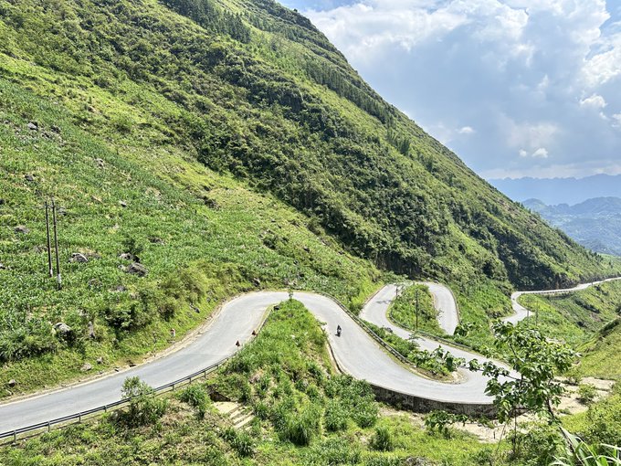 A scenic view of a winding mountain road with multiple switchbacks cutting through a lush green hillside under a partly cloudy sky.
