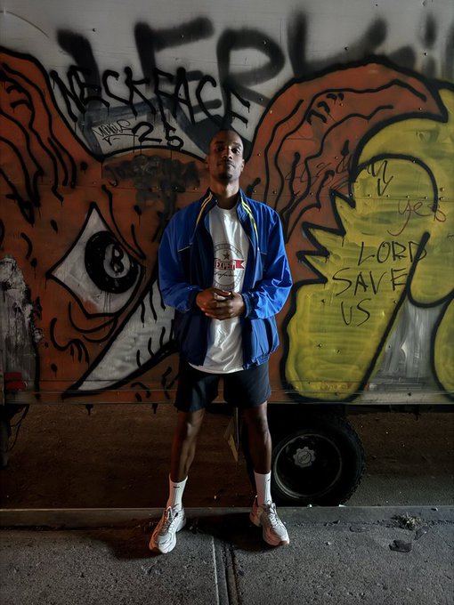 A man in a blue jacket, white t-shirt, and black shorts standing in front of a graffiti-covered wall that includes an 8-ball and text reading 'LORD SAVE US'.