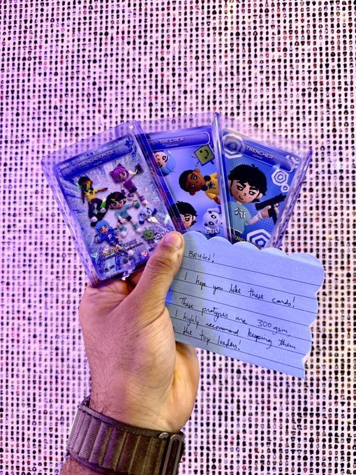 A hand holds three "Trencher TCG" trading cards alongside a handwritten note reading "Beeple! I hope you like these cards! These prototypes are 300gsm. I highly recommend keeping them free top loaders!", displayed against a shimmering sequined background.