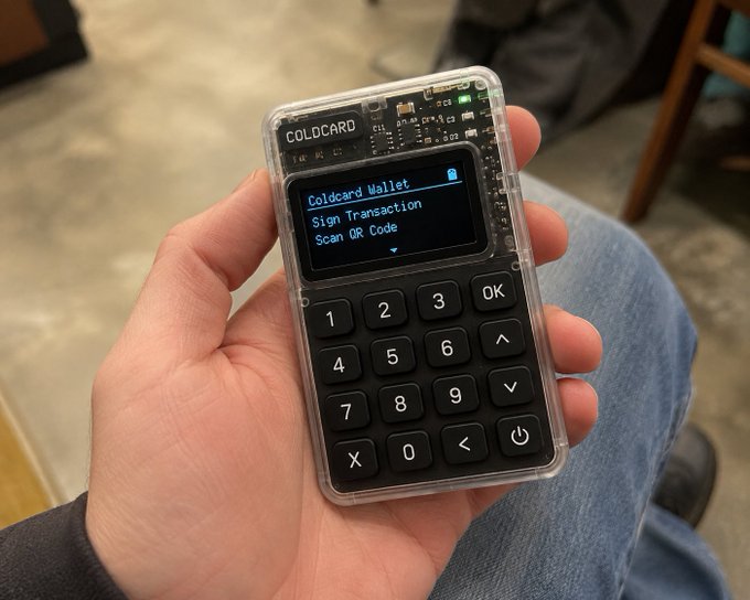 A hand holds a Coldcard hardware wallet displaying "Coldcard Wallet," "Sign Transaction," and "Scan QR Code" options on its screen, directly illustrating the security vulnerability referenced in the post about a 5-year-old bug that led to $1.6 million in Bitcoin being stolen.