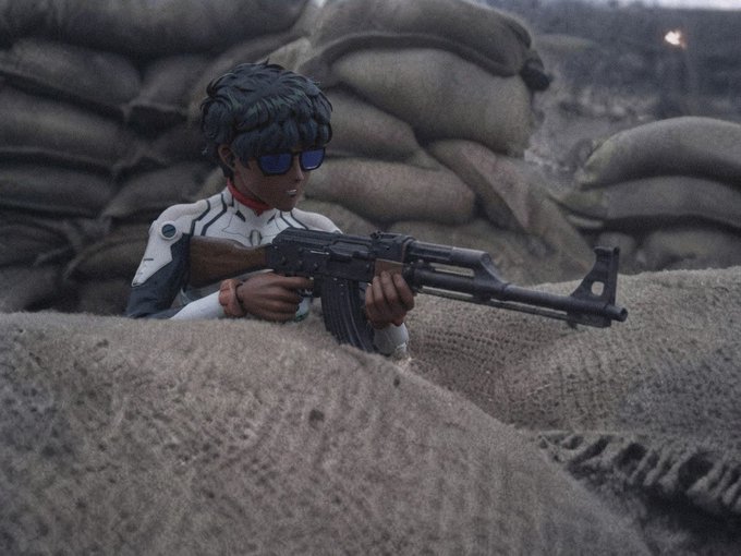 A stylized 3D-rendered character with dark hair and blue sunglasses aims an AK-47 rifle from behind sandbag bunkers in a war scene.