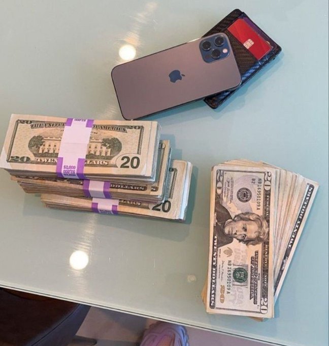 Stacks of $20 bills bound with $5,000 currency straps sit on a glass table next to a $20 bill fanned out (with $5,000, $10,000, and $20 denominations visible) and an iPhone in a carbon fiber case. The image visually represents the "grinding for the money" hustle by displaying the cash earned alongside the phone, likely used for work or business.