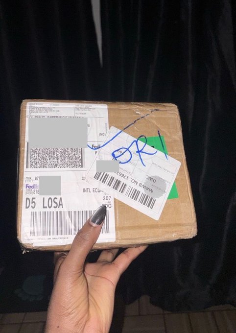 A hand holds a small shipping parcel covered with FedEx-style shipping labels, barcodes, handwritten blue markings, and redacted address information.