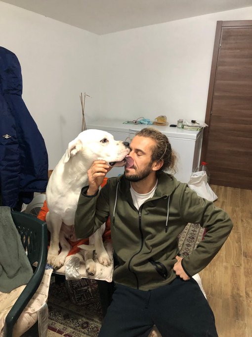 A casual indoor photo of a man with curly hair sitting next to a white dog that is licking his face, in a simply furnished room.