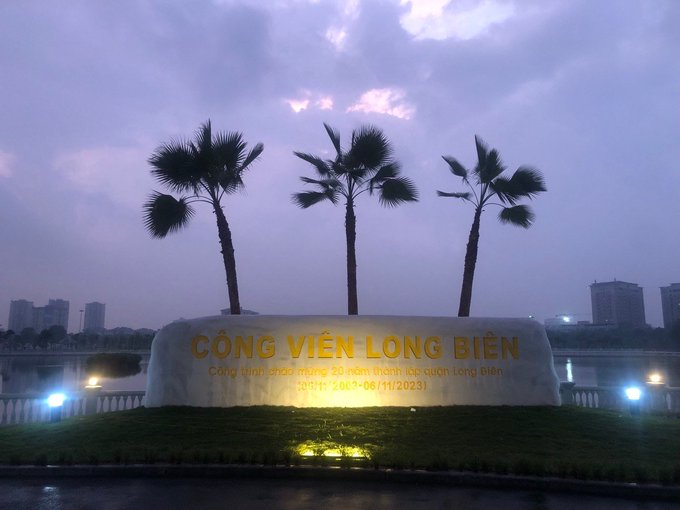 The image features a large white sign with three palm trees positioned behind it, creating an interesting visual effect against the backdrop of a city skyline at dusk. The sign displays the text "Cong Vien Long Bien" which translates to "Welcome to Long Bien". The scene is captured from a low angle, making the palm trees appear larger and more prominent in the image.