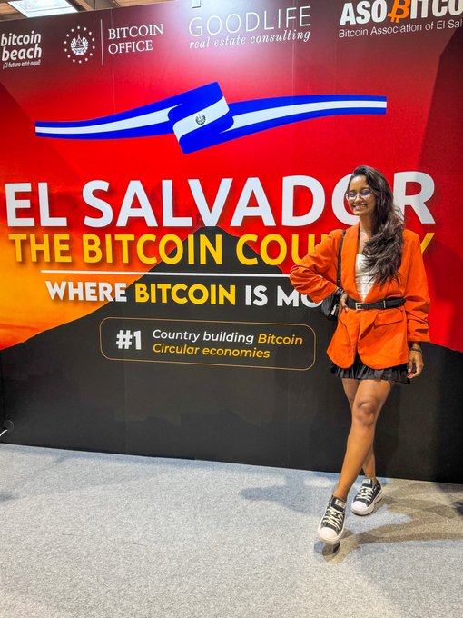 The image features a woman standing next to a large red sign that reads "El Salvador The Bitcoin Country Building Bitcoin Countries Circular Economies". She is wearing an orange jacket, black skirt, and white sneakers. The background of the photo shows a booth at a convention or event related to Bitcoin.