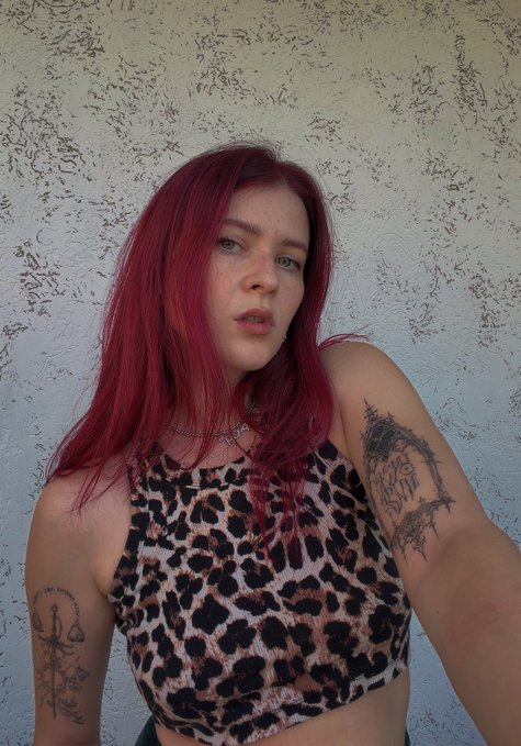 A selfie of a woman with vibrant red hair and freckles, wearing a leopard-print crop top and silver necklaces, with tattoos visible on her arms against a textured white wall.