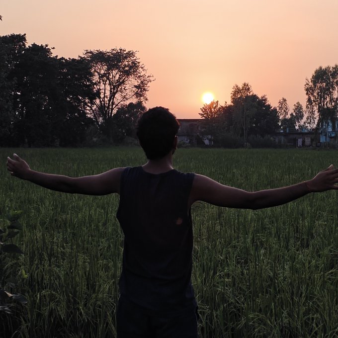 A silhouetted figure with outstretched arms stands in a grassy field, watching the sun set behind a distant tree line.