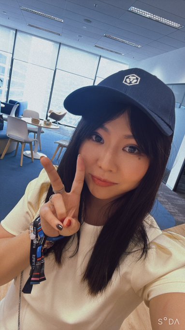 A selfie of a woman wearing a black cap with a logo and a beige top, making a peace sign in a modern office or cafe setting with large windows, watermarked 'S*DA'.