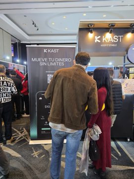 Two people stand before a booth at what appears to be a KAST event, with a banner reading 'MUEVE TU DINERO SIN LIMITES' and 'KAST KEEP IT SIMPLE' visible.