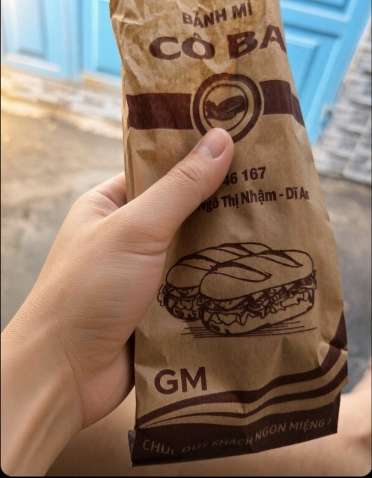 A hand holding a brown paper bag labeled “BÁNH MÌ CÔ BA” with a sandwich illustration.