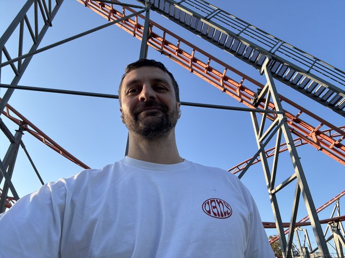 A selfie of a bearded man in a white sweatshirt with a 'WEEX' logo, taken beneath a roller coaster structure against a blue sky.