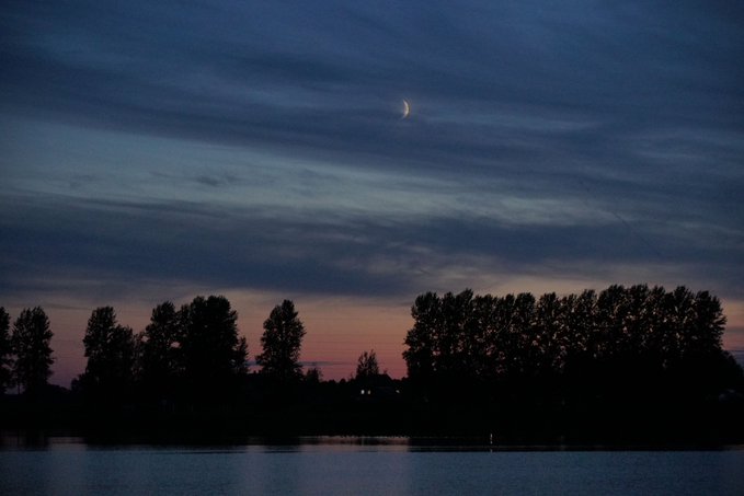 A serene twilight scene captures a crescent moon hanging in a deep blue sky streaked with soft clouds, above a silhouetted treeline bordering a calm body of water. The last glow of sunset paints the horizon in faint pink and orange hues, evoking the peaceful end of the day that perfectly complements the "Good night fren" sentiment, wishing viewers a tranquil night's rest under the fading light.