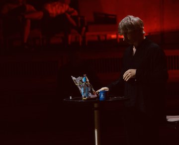 The image shows a man standing at a table with a laptop open, possibly working or browsing the internet. The background features a red wall, which could be part of an event like Web3 Summit happening in the room. There are also two chairs visible in the scene, one near the left side and another on the right side of the image.
