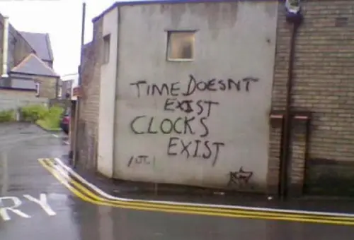 A building corner with boarded-up windows features graffiti spray-painted with the philosophical message 'TIME DOESN'T EXIST CLOCKS EXIST'.