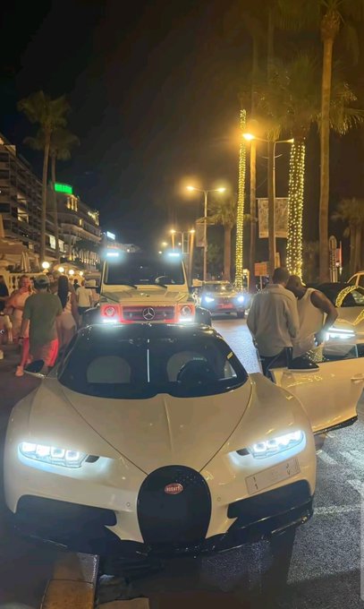 A white Bugatti Chiron supercar with illuminated headlights parked on a street at night in what appears to be a Mediterranean resort area, with palm trees decorated with string lights and a Mercedes G-Wagon visible in the background.