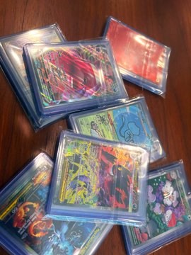 A collection of holographic trading cards, appearing to be Yu-Gi-Oh cards, scattered on a wooden surface in protective sleeves.