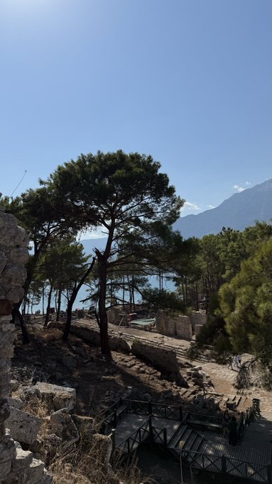Scenic coastal view with pine trees in the foreground, ancient stone ruins of a theater or amphitheater carved into the hillside, and the sea and mountains visible in the background under a clear blue sky.