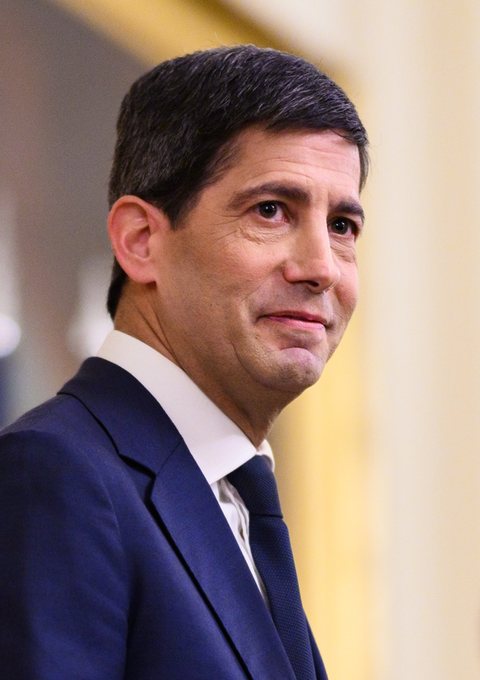 The image features a man dressed in a suit with a tie looking up at something off-camera. The background appears to be a building or room within an office setting. Above him, a wall can be seen, providing context for his location. The text "The Fed will inject $6,576,000,000 of liquidity into the economy next week." is displayed on this wall above the man's head. This suggests that there may be some financial news or event happening which has caught the man's attention, and it could be relevant to the image as well.