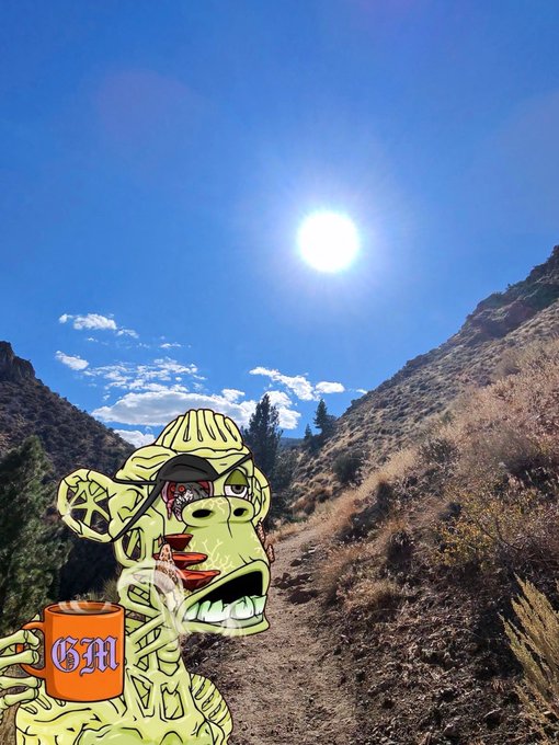 A cartoon-style ape character with a skull motif wearing a beanie, smoking and holding an orange 'GM' mug, set against a real photo background of a sunny desert mountain landscape.