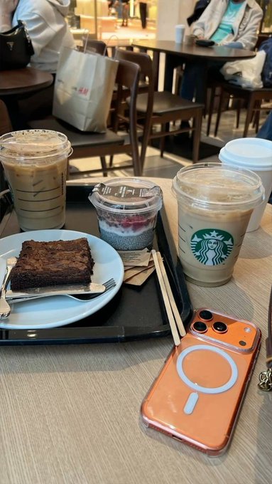A casual cafe table scene featuring two iced Starbucks drinks, a brownie on a white plate, a chia pudding, chopsticks, and an orange-cased iPhone with a magnetic ring.