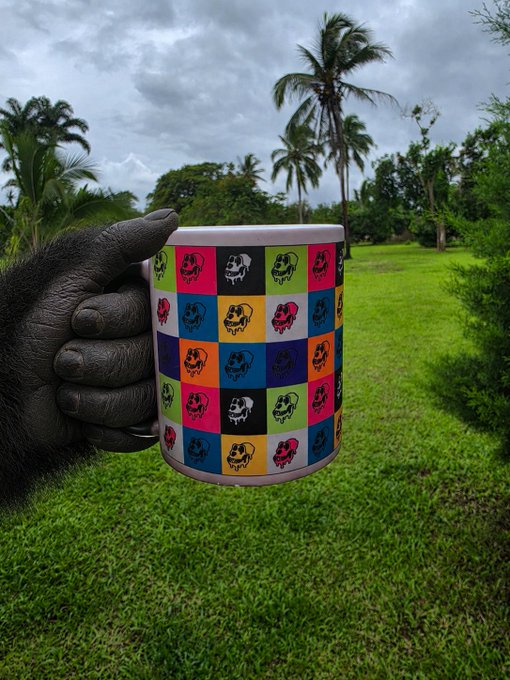 The image features a hand holding up a colorful mug with a picture of a face on it, which appears to be a cartoon or patterned design. The mug is being held above a grassy area that includes palm trees, suggesting an outdoor setting. The background of the photo also contains some text, but it does not provide any additional information about the image's context.