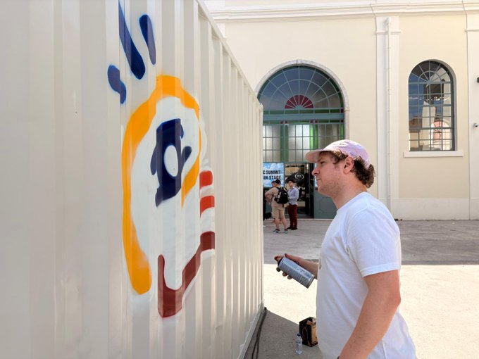 The image features a man standing next to a white building, spray painting graffiti on its side. The graffiti artwork includes an American flag design with a hand holding a paintbrush. The man appears to be wearing a white shirt as he works on his art project. In the background, there are other people present who might be observing or participating in the graffiti-making process.