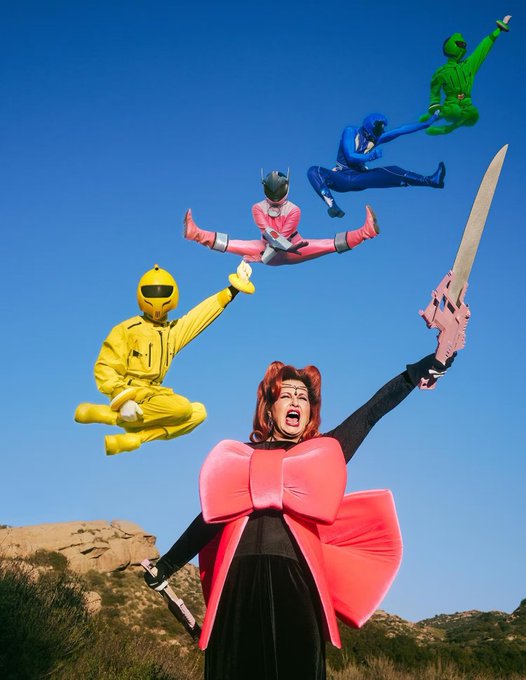 A person dressed as a Red Power Ranger with red hair stands triumphantly with a pink sword raised overhead against a clear blue sky, while four other Power Rangers in pink, yellow, blue, and green appear to leap and pose dynamically above the rocky desert landscape. The bold, heroic cosplay tableau reflects the post's rallying call for fellow "blockchain collector nerds" to join in playful fandom spirit on The God Pull platform.