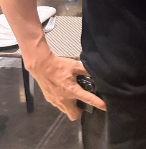 A muscular forearm with prominently visible veins grips a smartphone near the hip of someone wearing dark clothing, with a textured gray rug visible in the background. The image appears to be a casual up-close shot of a person's hand and phone, seemingly shared in response to the flattering comment about prominent hand veins.